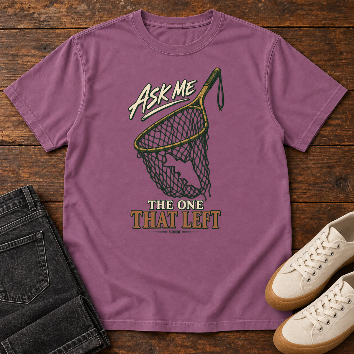 Berry Ask Me About the One That Left T-Shirt with an empty fishing landing net and cream-and-copper lettering, front flat lay on wood.