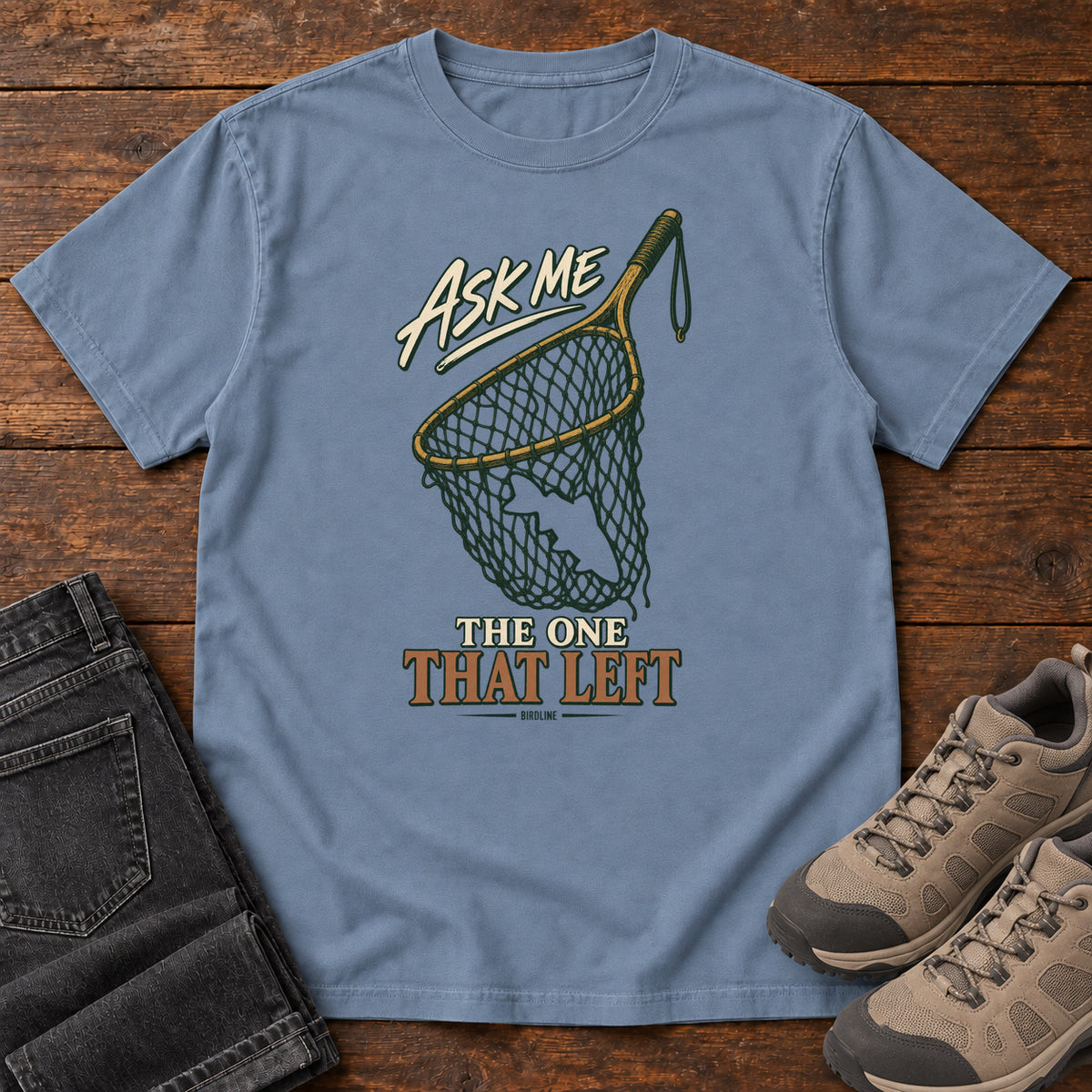 Blue Jean Ask Me About the One That Left T-Shirt with an empty fishing landing net and cream-and-copper lettering, front flat lay on wood.