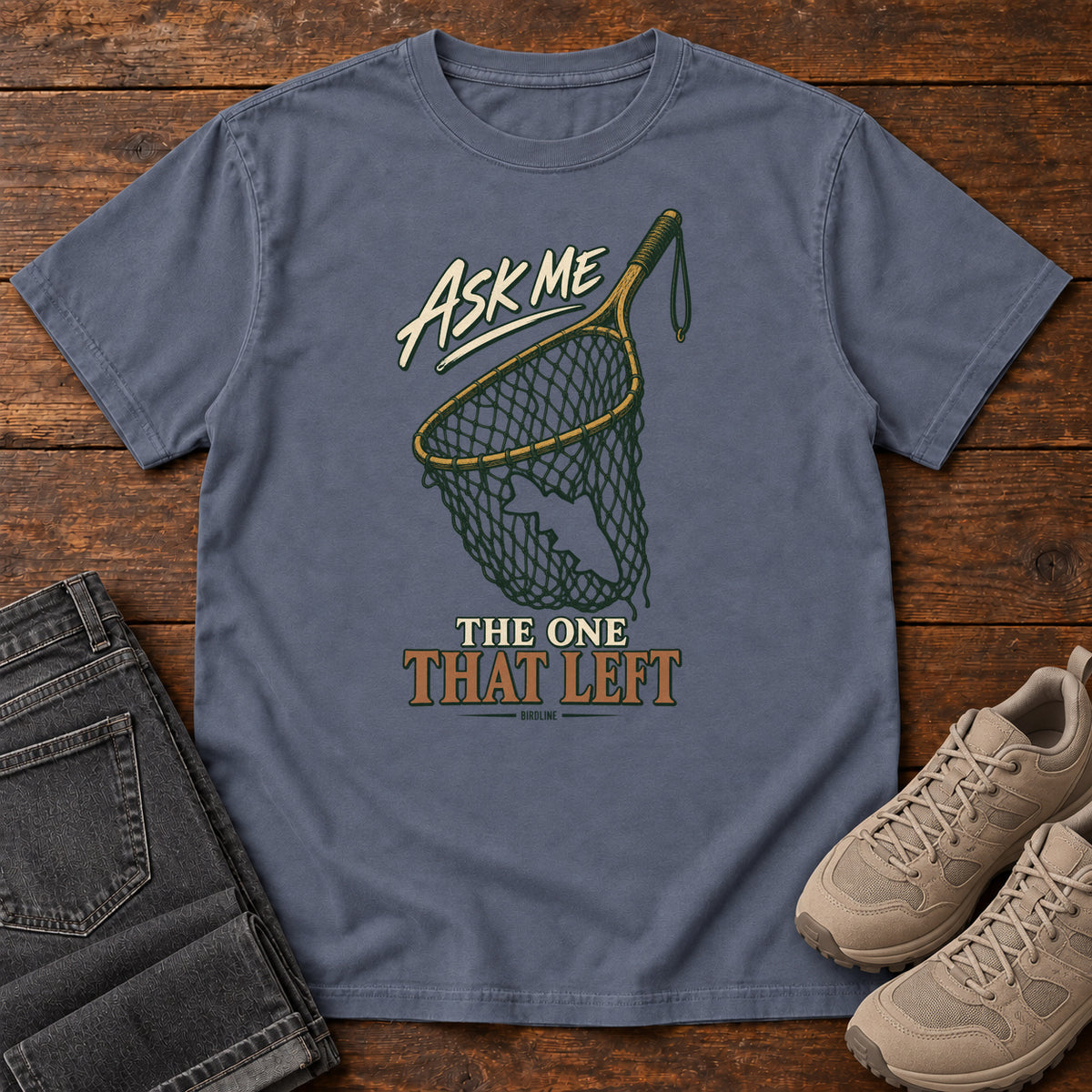 Denim Ask Me About the One That Left T-Shirt with an empty fishing landing net and cream-and-copper lettering, front flat lay on wood.