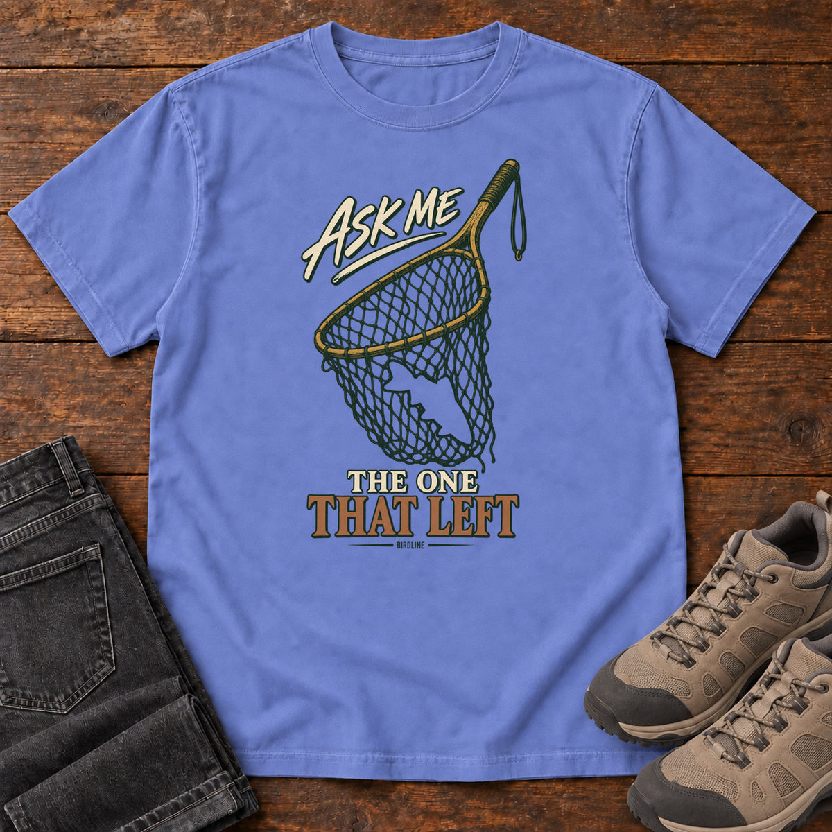 Flo Blue Ask Me About the One That Left T-Shirt with an empty fishing landing net and cream-and-copper lettering, front flat lay on wood.