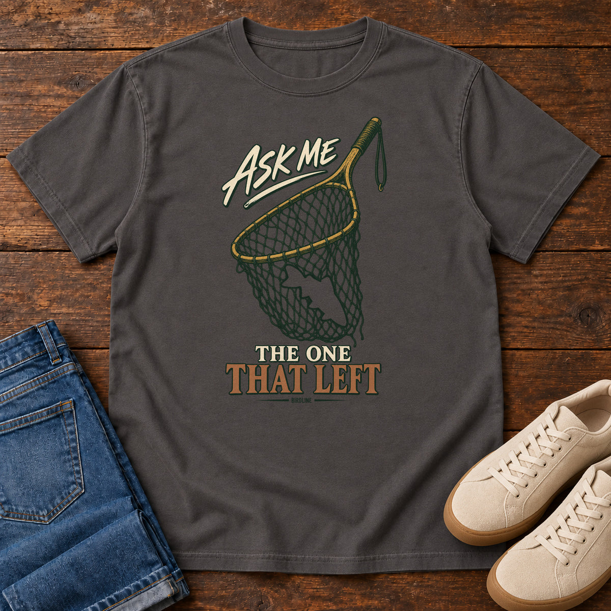 Graphite Ask Me About the One That Left T-Shirt with an empty fishing landing net and cream-and-copper lettering, front flat lay on wood.