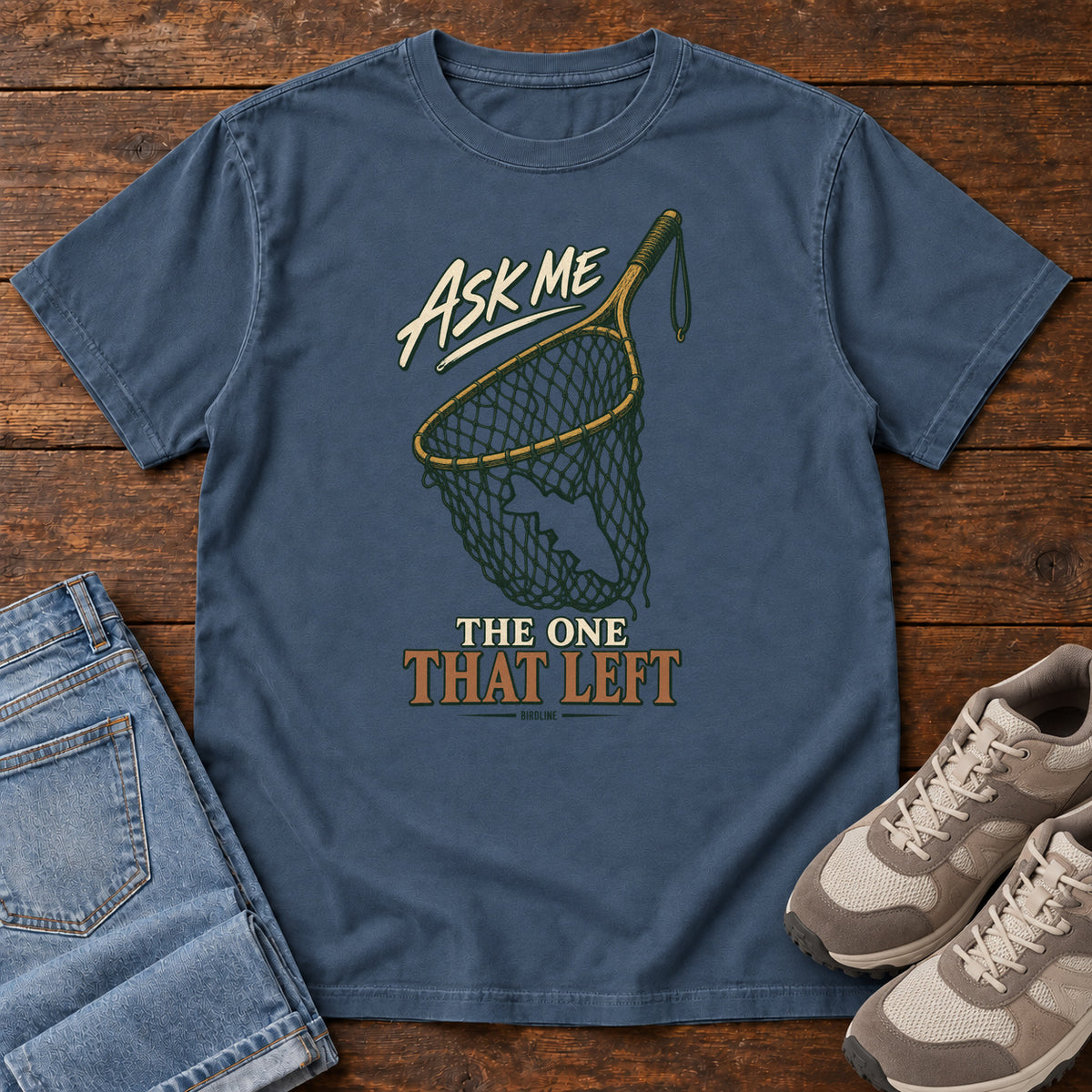Midnight Ask Me About the One That Left T-Shirt with an empty fishing landing net and cream-and-copper lettering, front flat lay on wood.