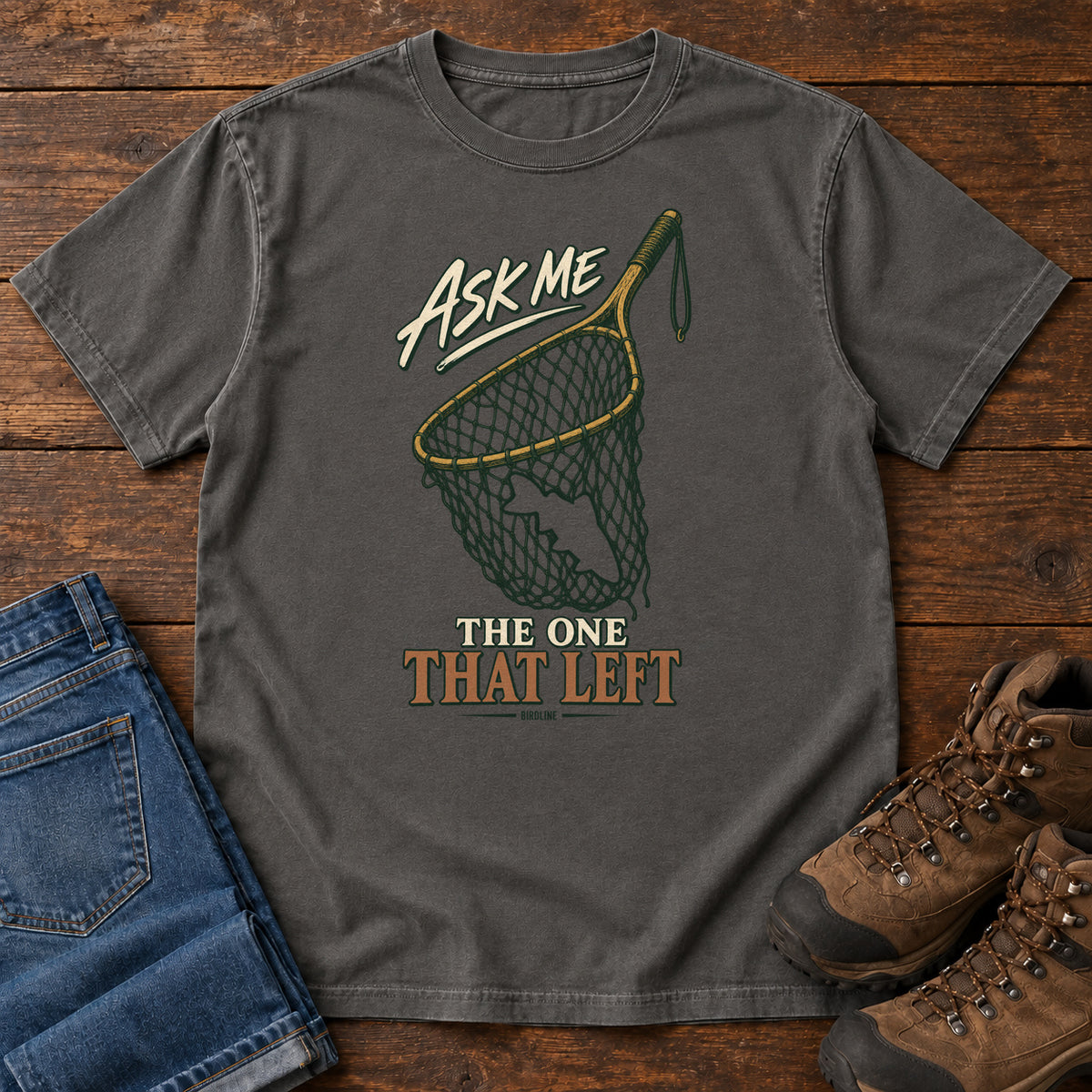 Pepper Ask Me About the One That Left T-Shirt with an empty fishing landing net and cream-and-copper lettering, front flat lay on wood.