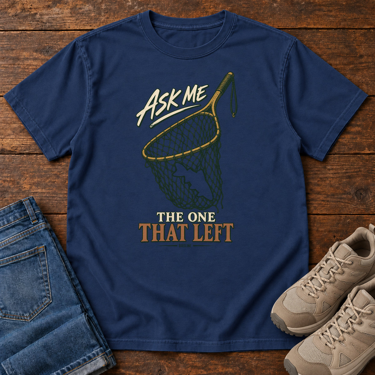 True Navy Ask Me About the One That Left T-Shirt with an empty fishing landing net and cream-and-copper lettering, front flat lay on wood.