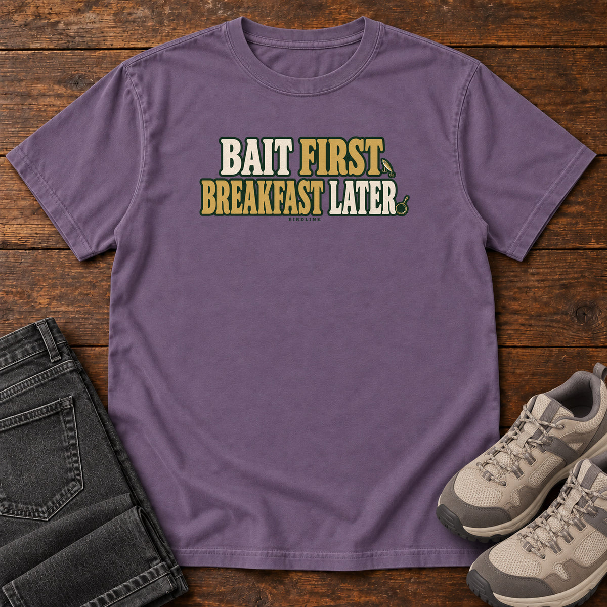 Grape Bait First Breakfast Later T-Shirt with cream and gold Bait First Breakfast Later lettering; front flat lay on wood.