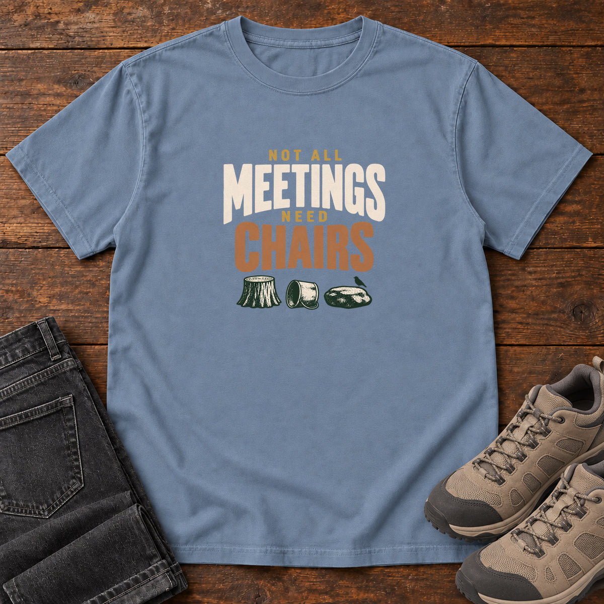 Blue Jean Not All Meetings Need Chairs T-Shirt with stacked cream-and-orange lettering above a stump, tipped mug and rock; front flat lay on wood.