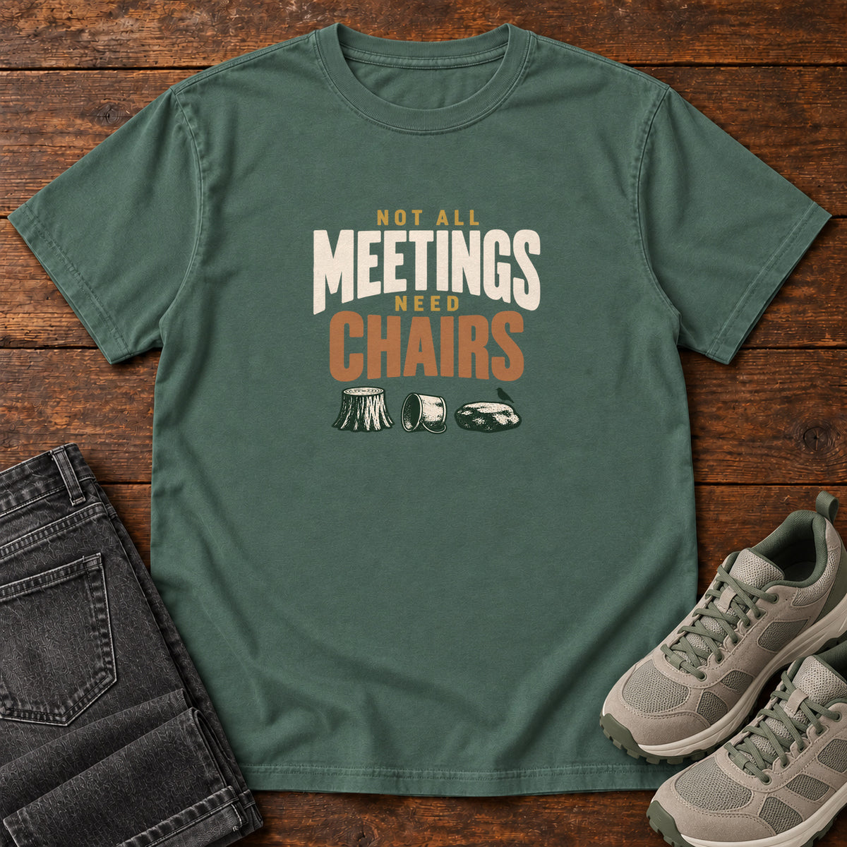 Blue Spruce Not All Meetings Need Chairs T-Shirt with stacked cream-and-orange lettering above a stump, tipped mug and rock; front flat lay on wood.