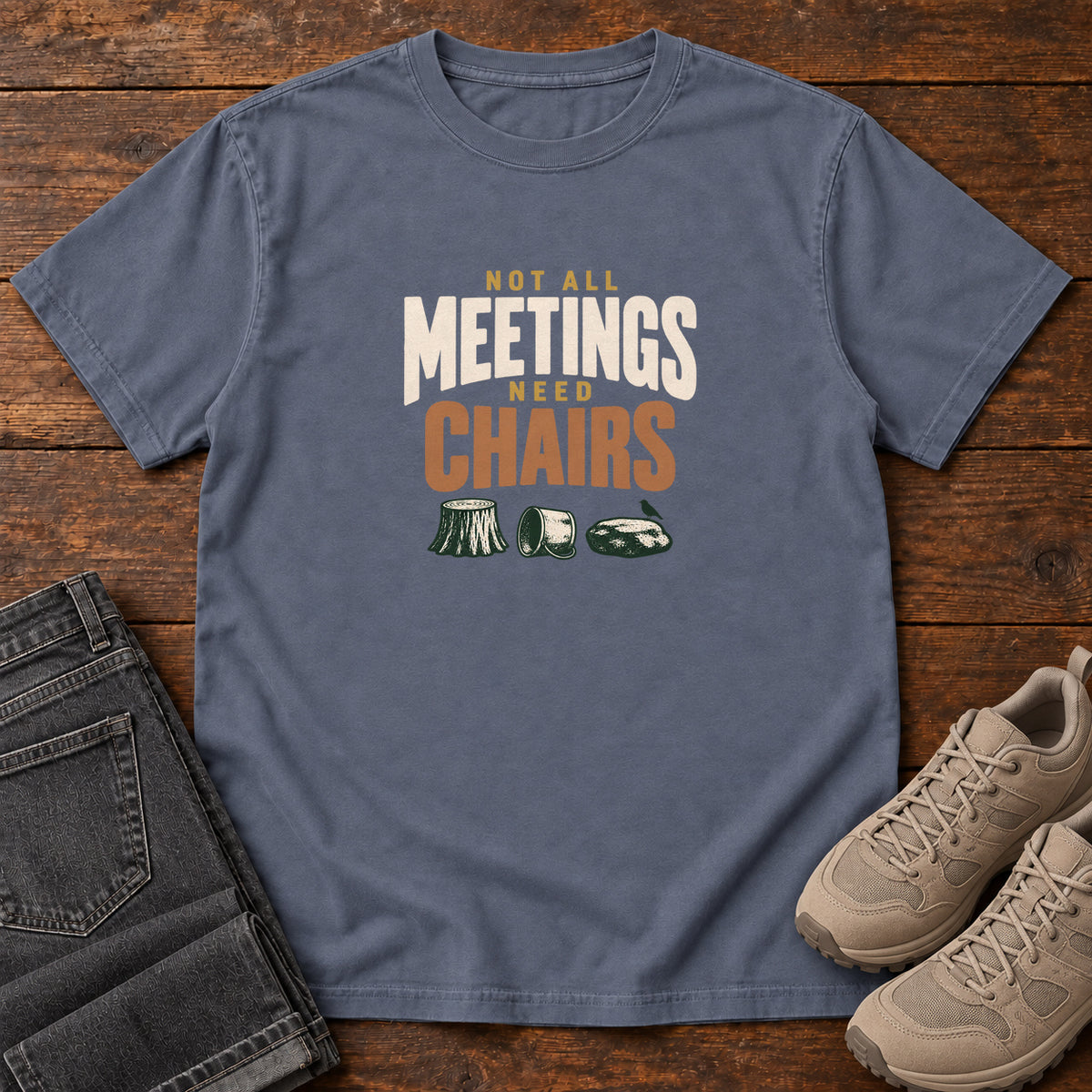 Denim Not All Meetings Need Chairs T-Shirt with stacked cream-and-orange lettering above a stump, tipped mug and rock; front flat lay on wood.