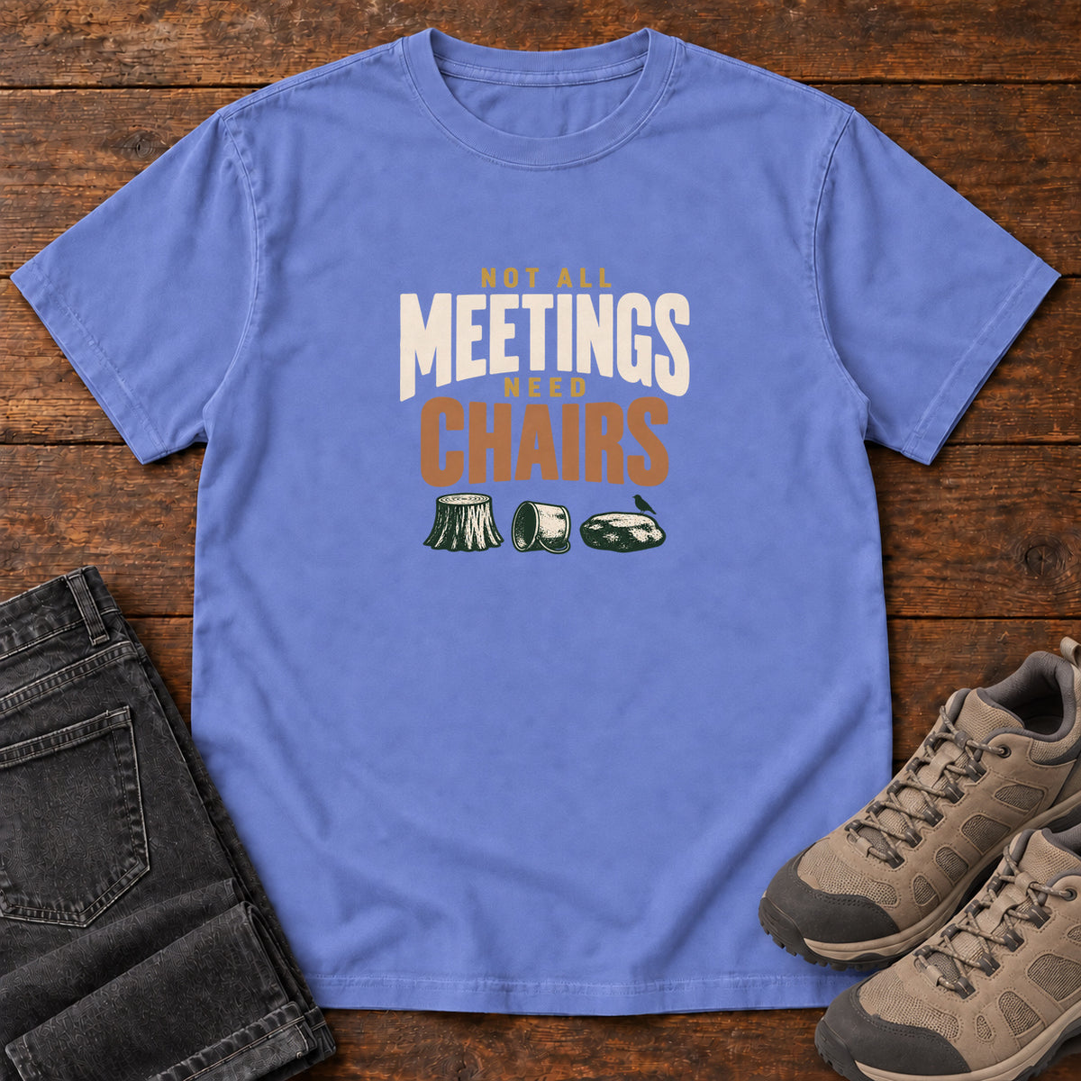 Flo Blue Not All Meetings Need Chairs T-Shirt with stacked cream-and-orange lettering above a stump, tipped mug and rock; front flat lay on wood.