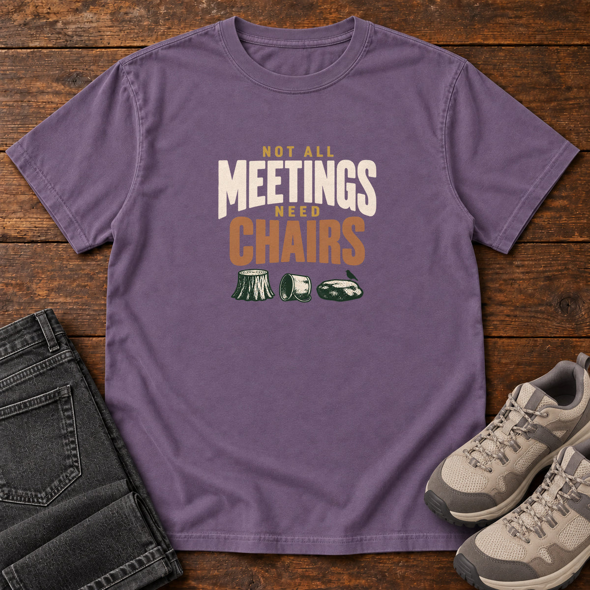 Grape Not All Meetings Need Chairs T-Shirt with stacked cream-and-orange lettering above a stump, tipped mug and rock; front flat lay on wood.