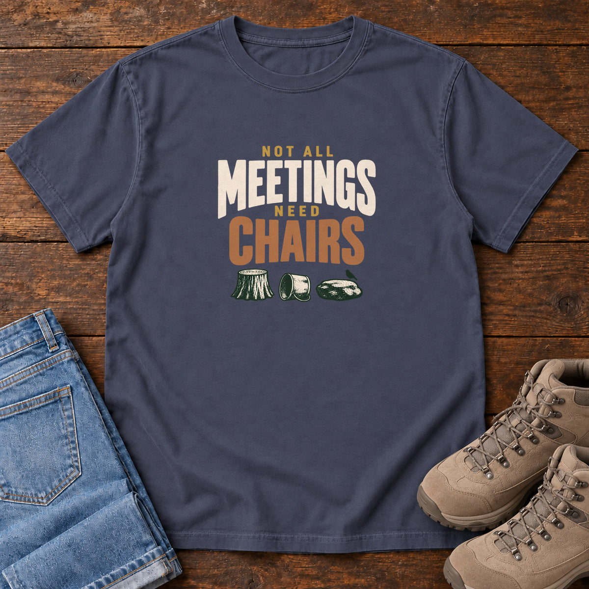 Navy Not All Meetings Need Chairs T-Shirt with stacked cream-and-orange lettering above a stump, tipped mug and rock; front flat lay on wood.