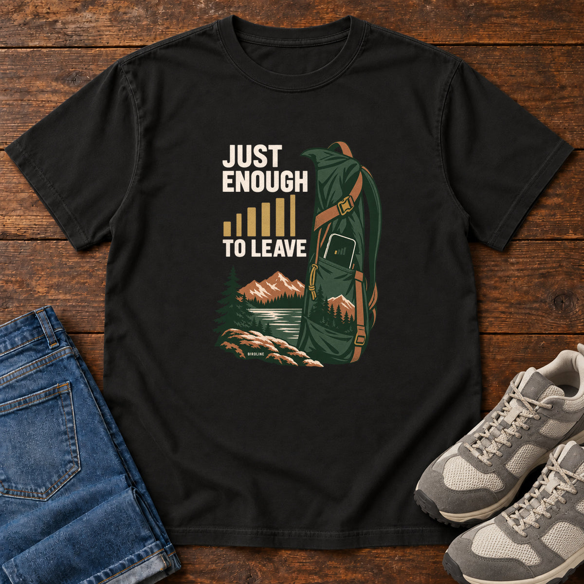 Black Just Enough Signal to Leave T-Shirt with a green hiking backpack, mountains and gold signal bars beside cream lettering, front flat lay on wood.