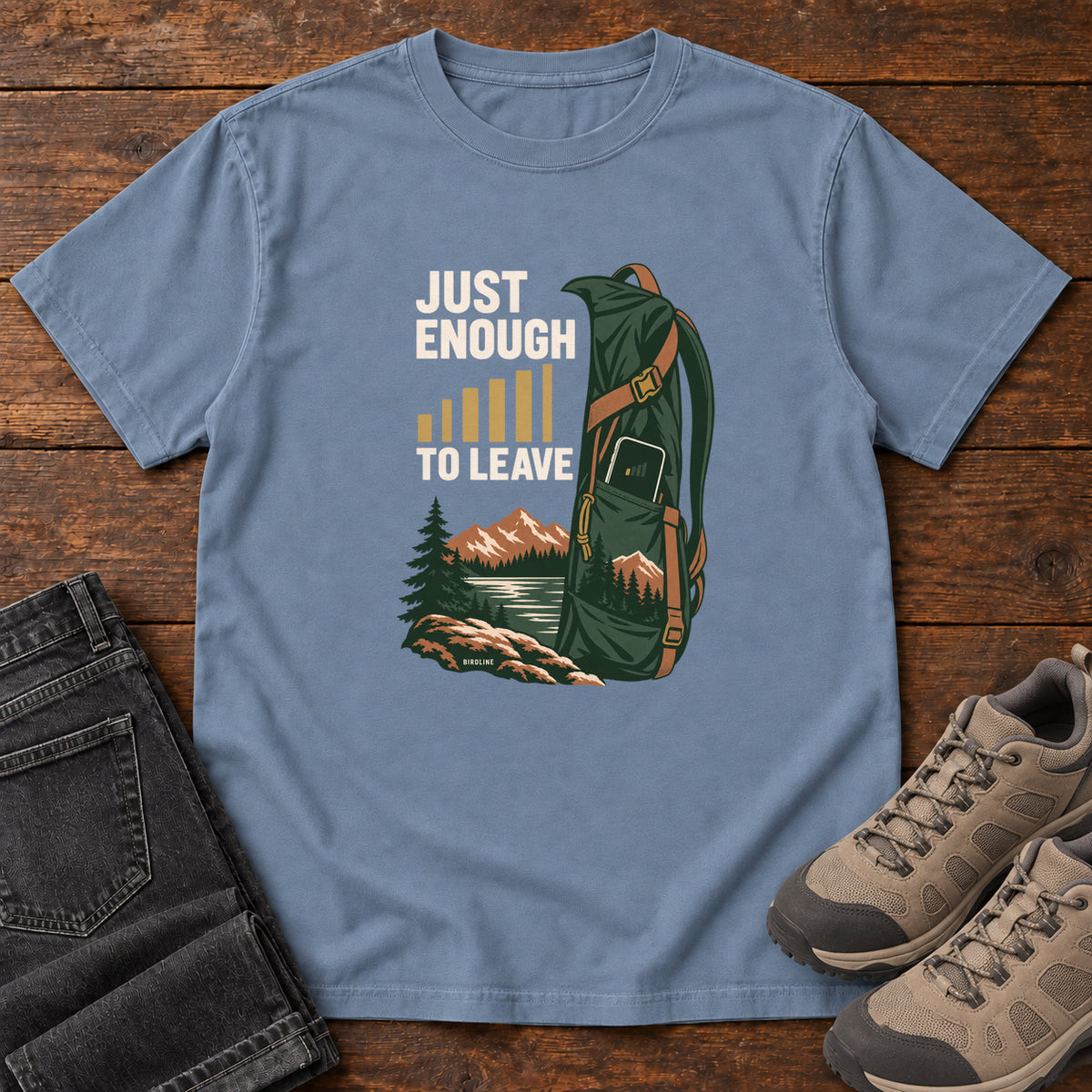 Blue Jean Just Enough Signal to Leave T-Shirt with a green hiking backpack, mountains and gold signal bars beside cream lettering, front flat lay on wood.