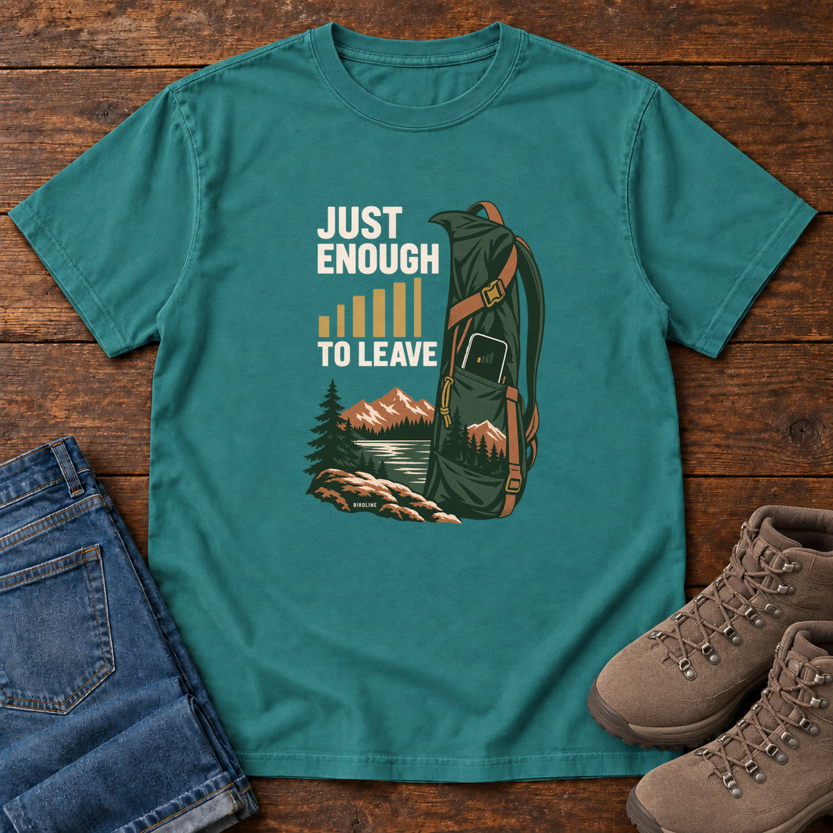 Emerald Just Enough Signal to Leave T-Shirt with a green hiking backpack, mountains and gold signal bars beside cream lettering, front flat lay on wood.