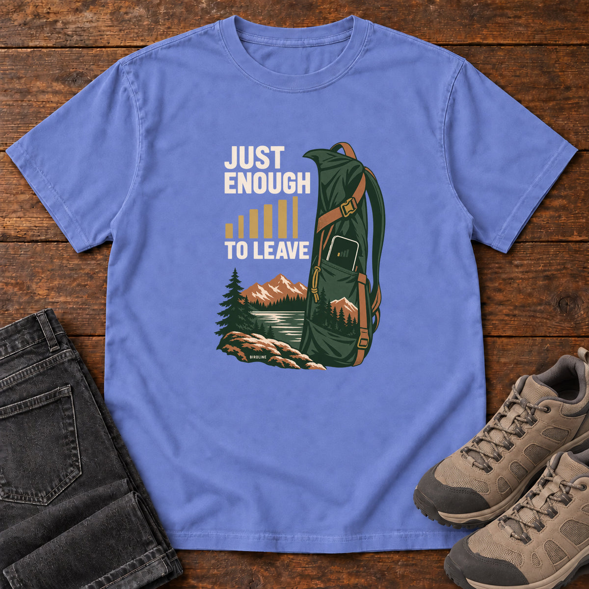 Flo Blue Just Enough Signal to Leave T-Shirt with a green hiking backpack, mountains and gold signal bars beside cream lettering, front flat lay on wood.