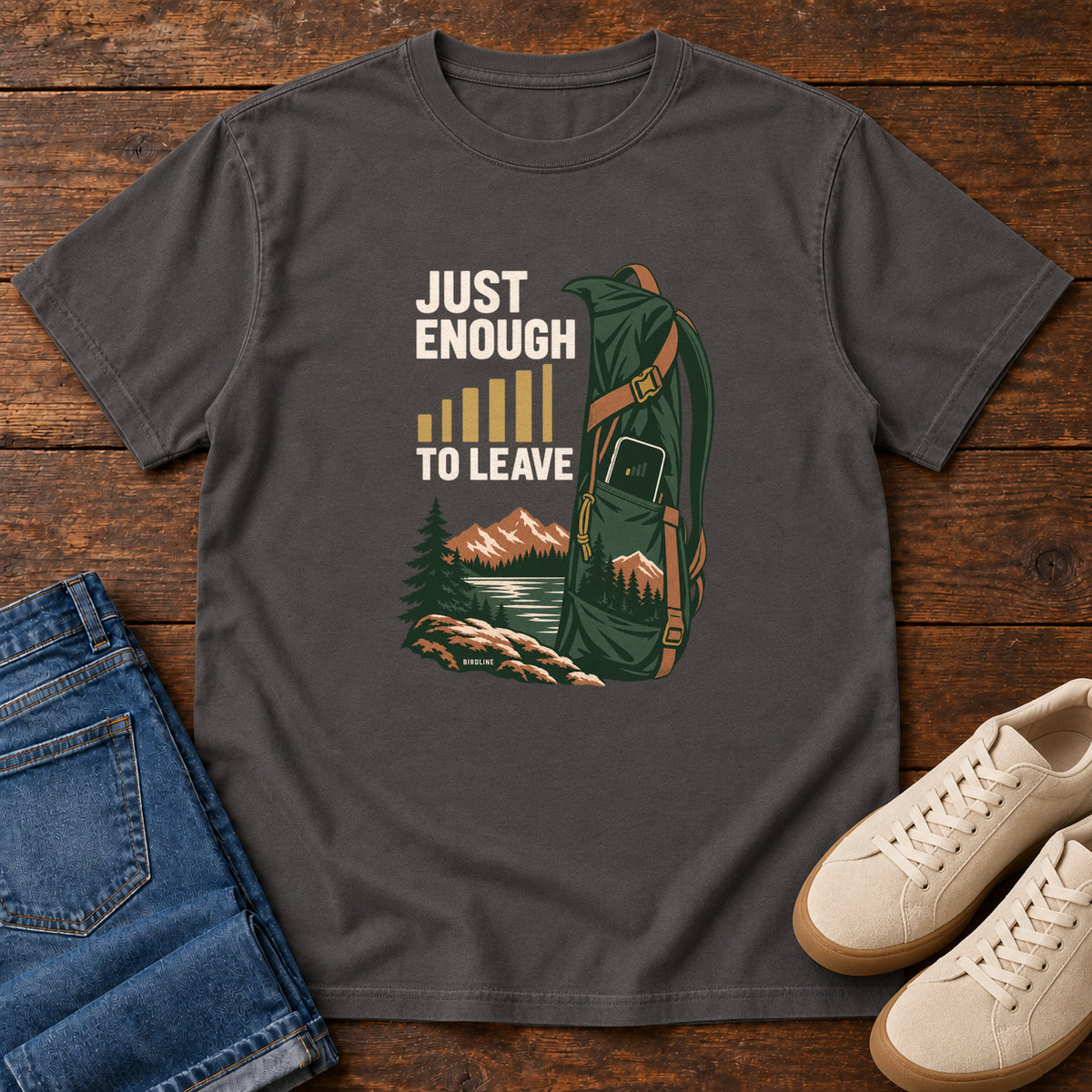 Graphite Just Enough Signal to Leave T-Shirt with a green hiking backpack, mountains and gold signal bars beside cream lettering, front flat lay on wood.