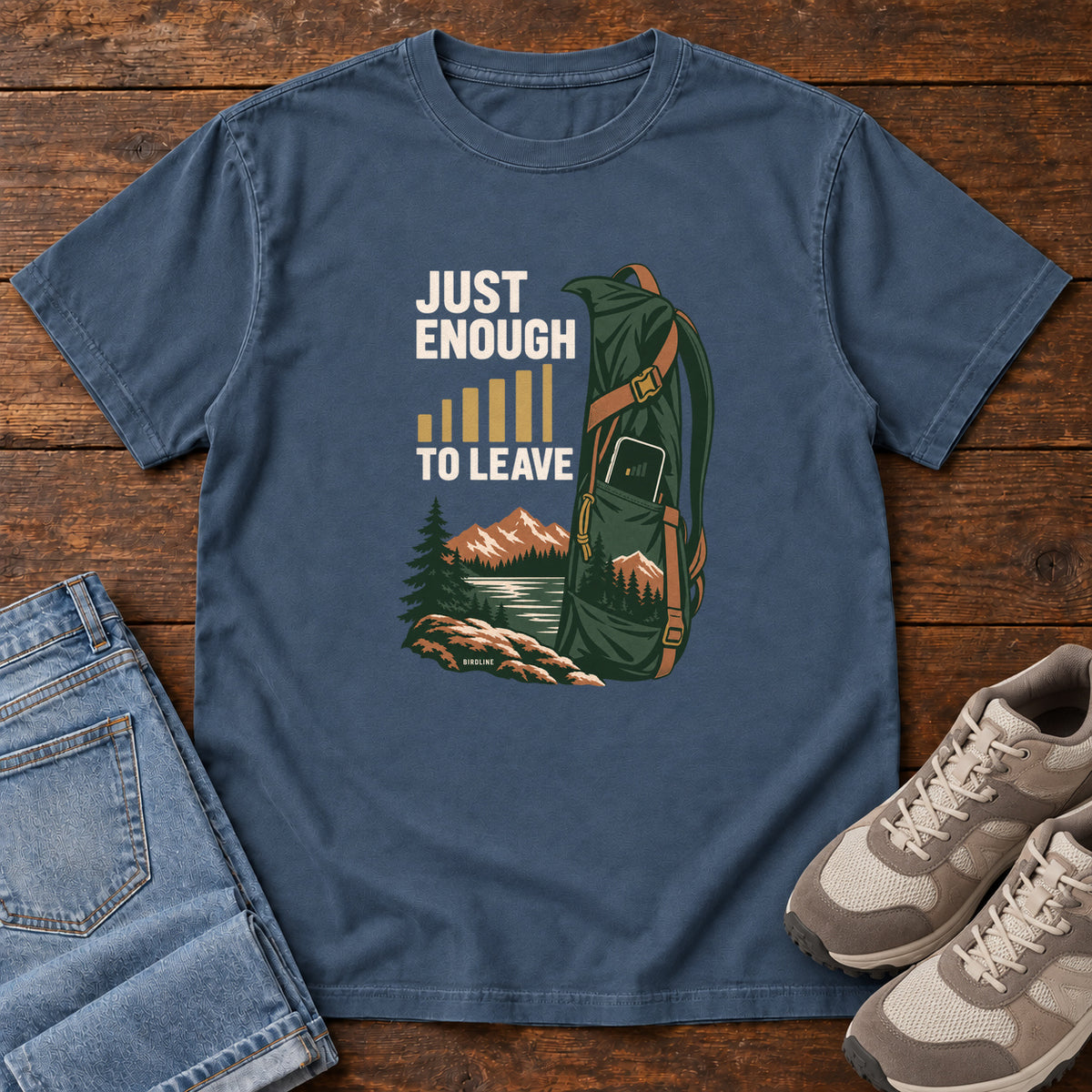 Midnight Just Enough Signal to Leave T-Shirt with a green hiking backpack, mountains and gold signal bars beside cream lettering, front flat lay on wood.