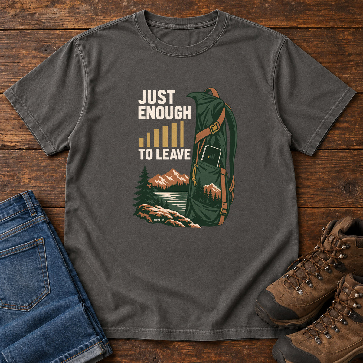 Pepper Just Enough Signal to Leave T-Shirt with a green hiking backpack, mountains and gold signal bars beside cream lettering, front flat lay on wood.