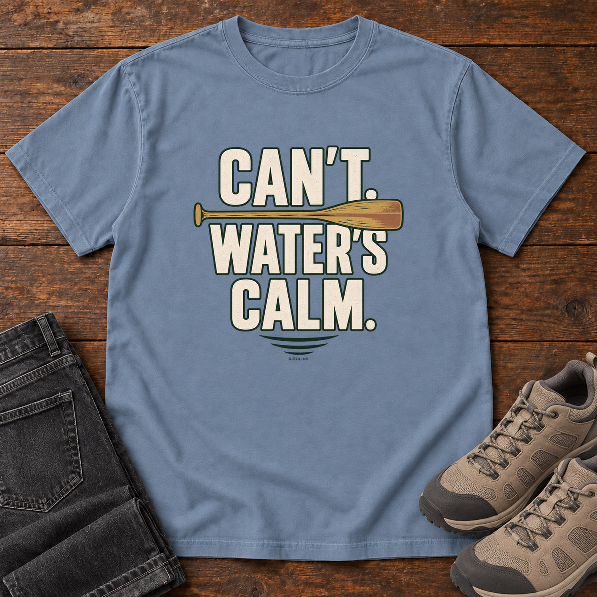 Can't. Water's Calm. T-Shirt in Blue Jean, front laid flat on wood, featuring bold cream slogan lettering divided by a horizontal orange paddle.