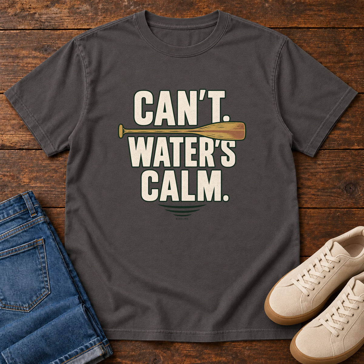 Can't. Water's Calm. T-Shirt in Graphite, front laid flat on wood, featuring bold cream slogan lettering divided by a horizontal orange paddle.