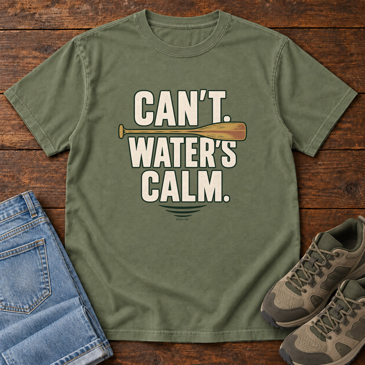 Can't. Water's Calm. T-Shirt in Moss, front laid flat on wood, featuring bold cream slogan lettering divided by a horizontal orange paddle.