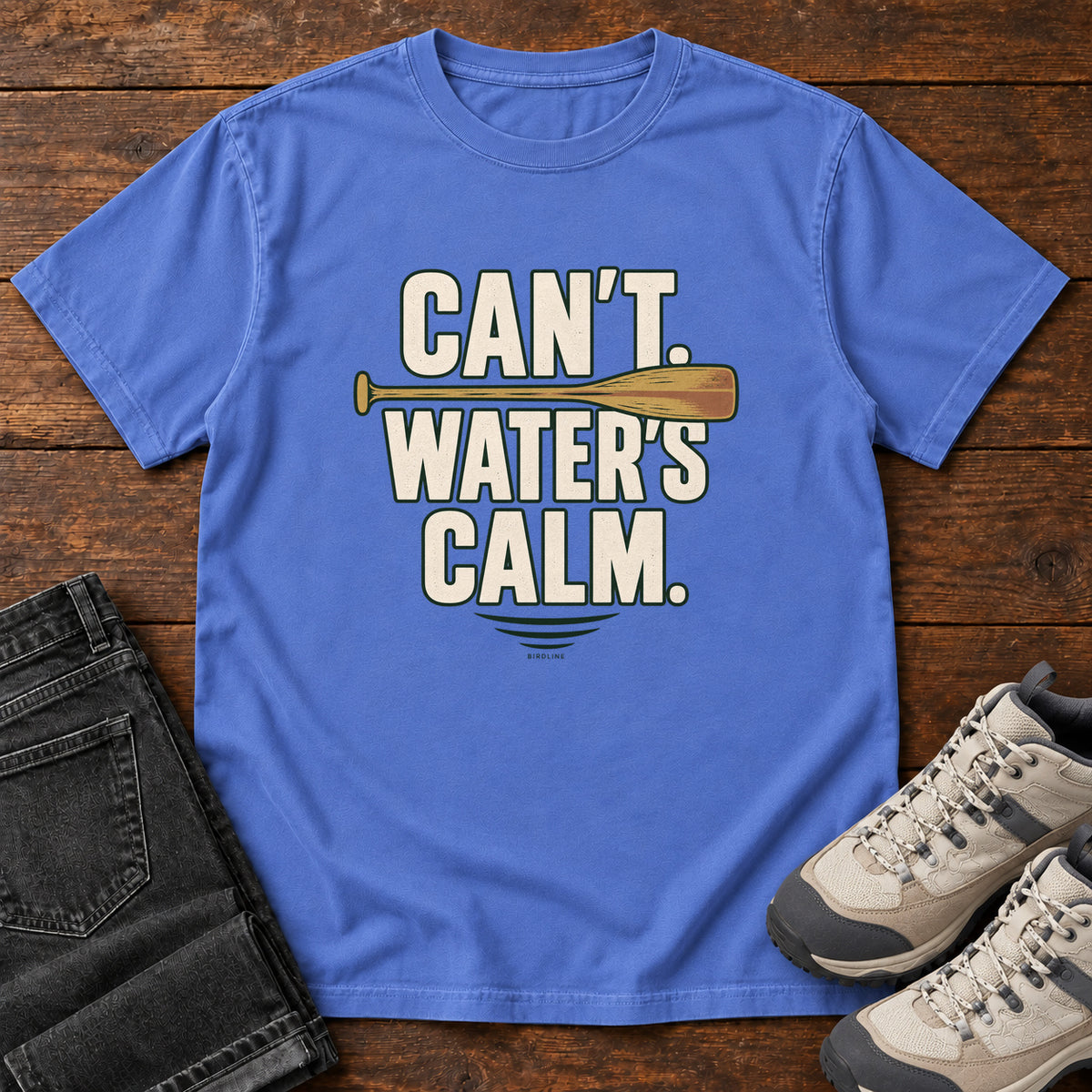 Can't. Water's Calm. T-Shirt in Mystic Blue, front laid flat on wood, featuring bold cream slogan lettering divided by a horizontal orange paddle.