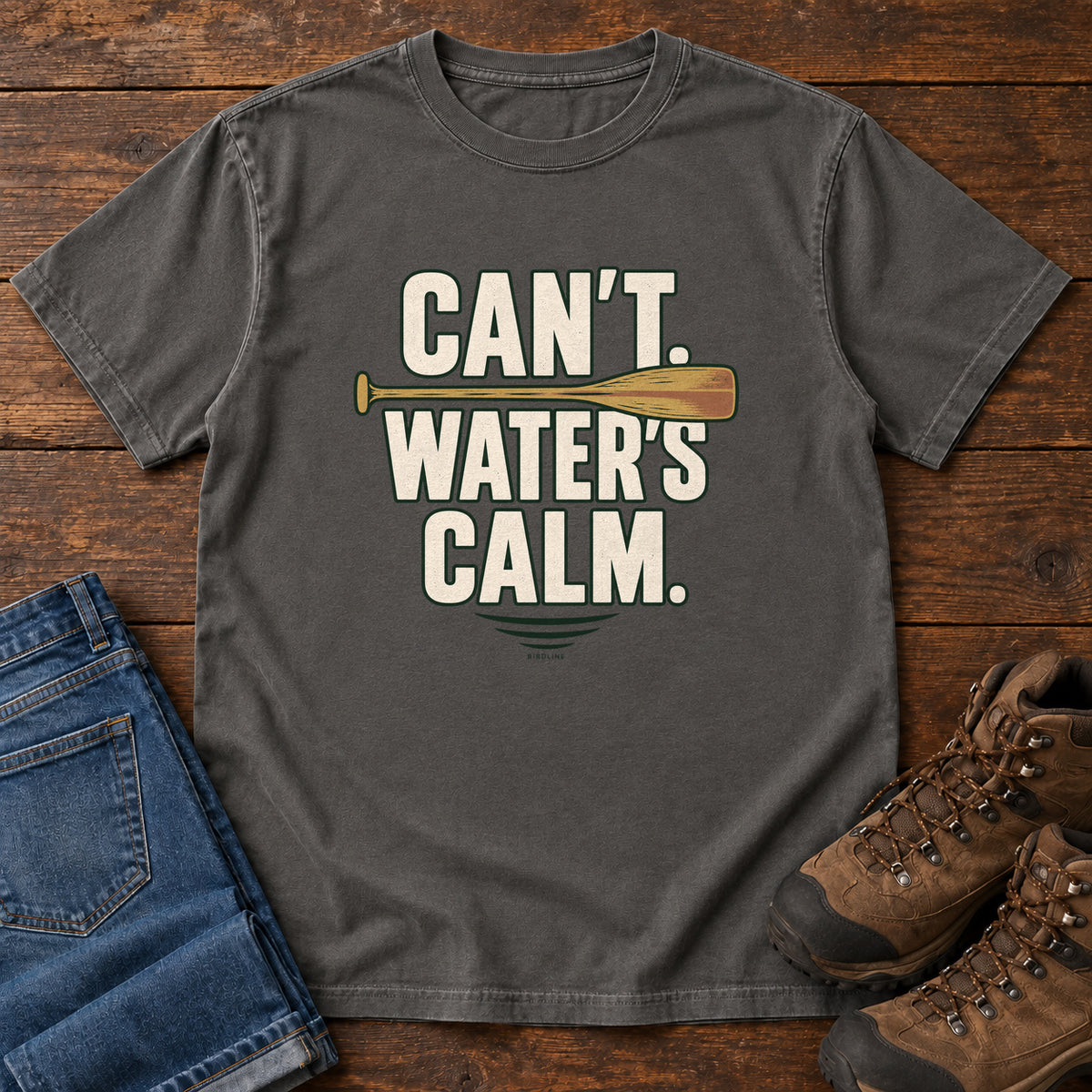 Can't. Water's Calm. T-Shirt in Pepper, front laid flat on wood, featuring bold cream slogan lettering divided by a horizontal orange paddle.