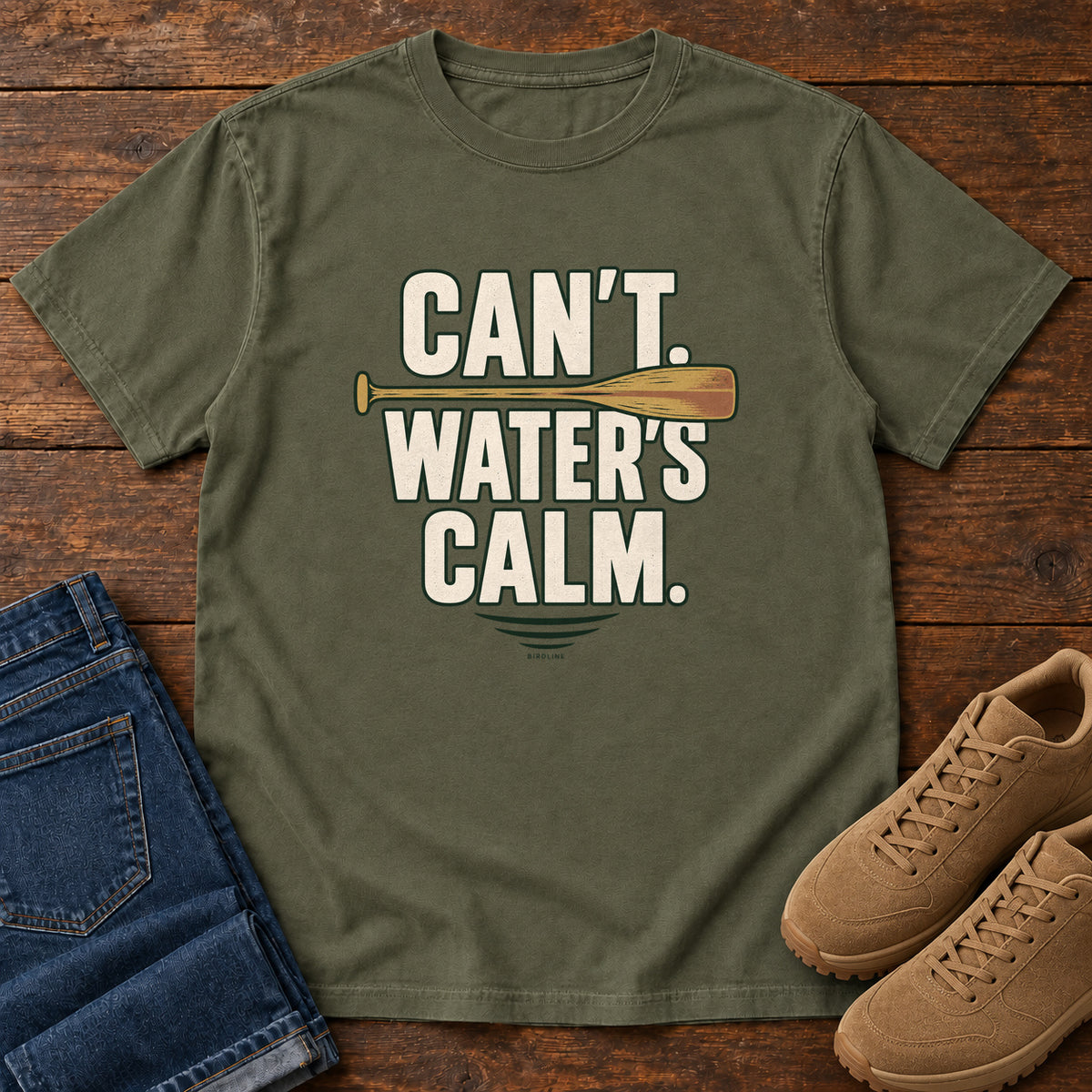Can't. Water's Calm. T-Shirt in Sage, front laid flat on wood, featuring bold cream slogan lettering divided by a horizontal orange paddle.