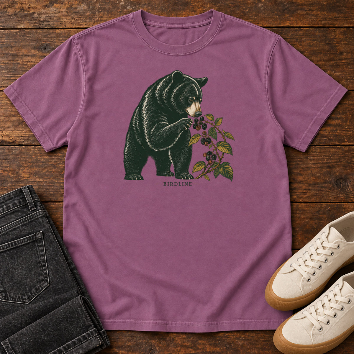 Berry Black Bear Berries Birdline T-Shirt with a dark bear holding a leafy berry branch, front flat lay on wood.