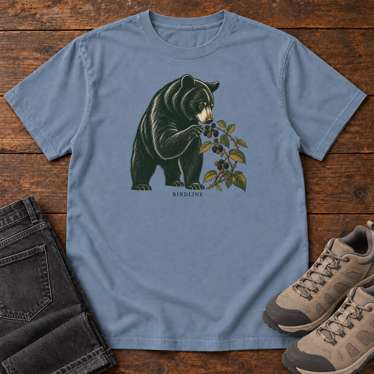 Blue Jean Black Bear Berries Birdline T-Shirt with a dark bear holding a leafy berry branch, front flat lay on wood.