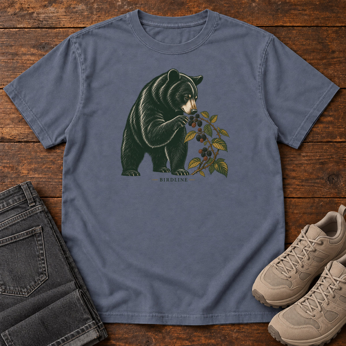Denim Black Bear Berries Birdline T-Shirt with a dark bear holding a leafy berry branch, front flat lay on wood.