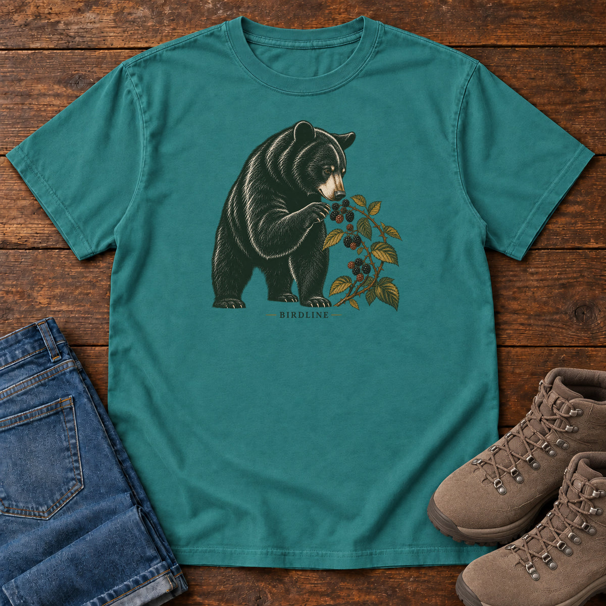 Emerald Black Bear Berries Birdline T-Shirt with a dark bear holding a leafy berry branch, front flat lay on wood.