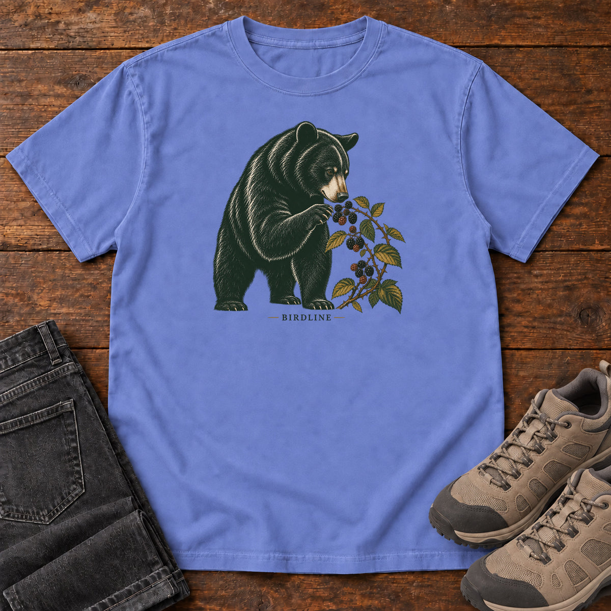 Flo Blue Black Bear Berries Birdline T-Shirt with a dark bear holding a leafy berry branch, front flat lay on wood.