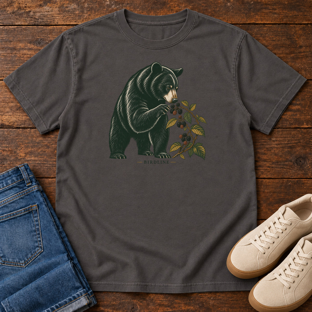 Graphite Black Bear Berries Birdline T-Shirt with a dark bear holding a leafy berry branch, front flat lay on wood.