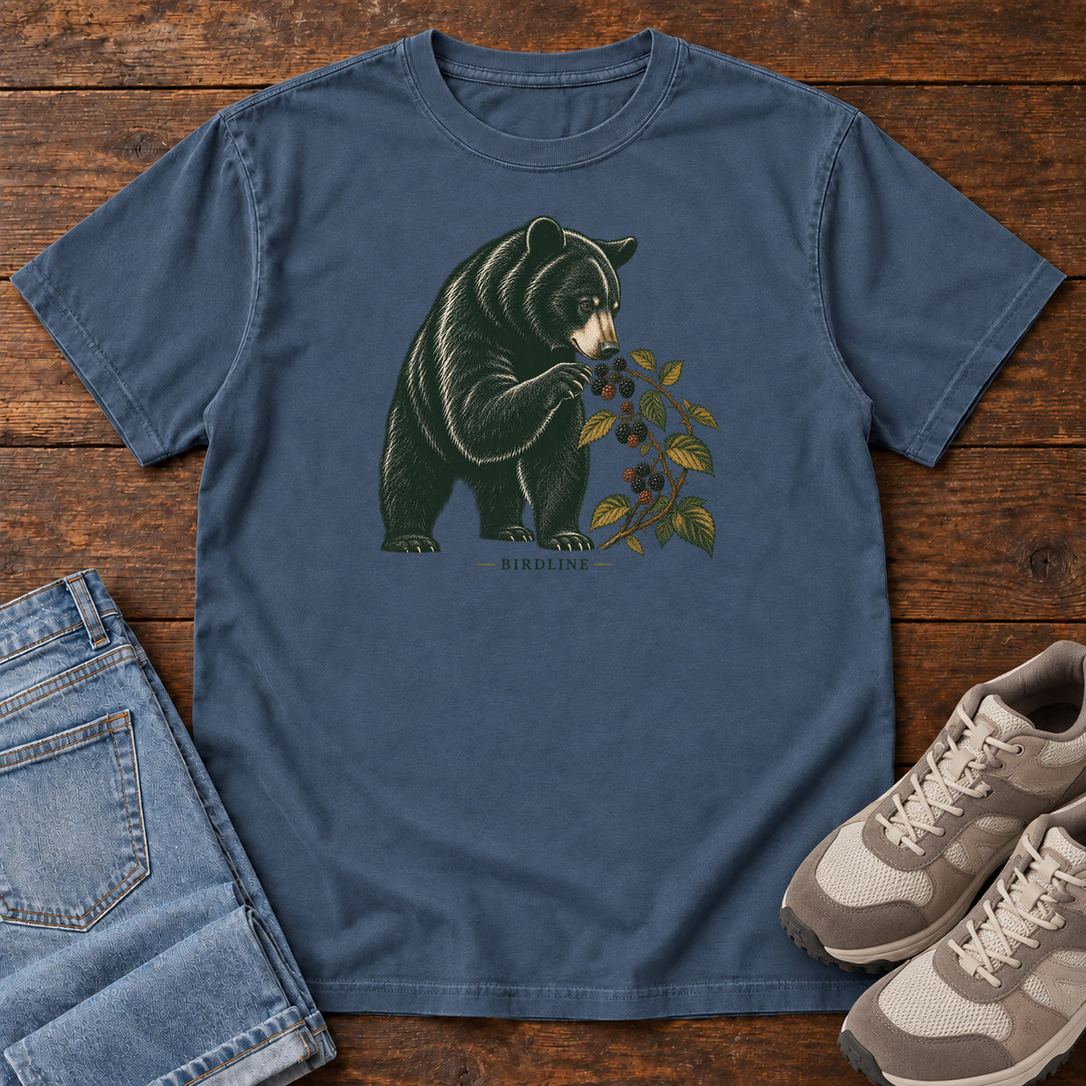 Midnight Black Bear Berries Birdline T-Shirt with a dark bear holding a leafy berry branch, front flat lay on wood.