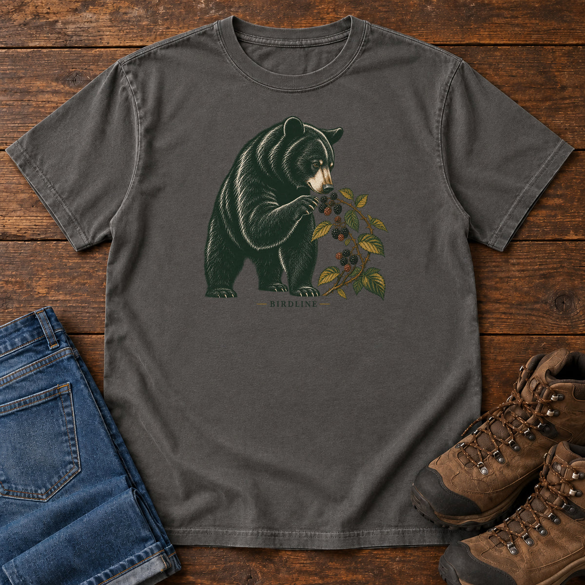 Pepper Black Bear Berries Birdline T-Shirt with a dark bear holding a leafy berry branch, front flat lay on wood.
