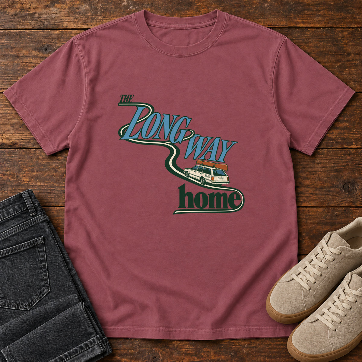 Brick The Long Way Home T-Shirt with a station wagon on a winding road woven through The Long Way Home lettering; front flat lay on wood.