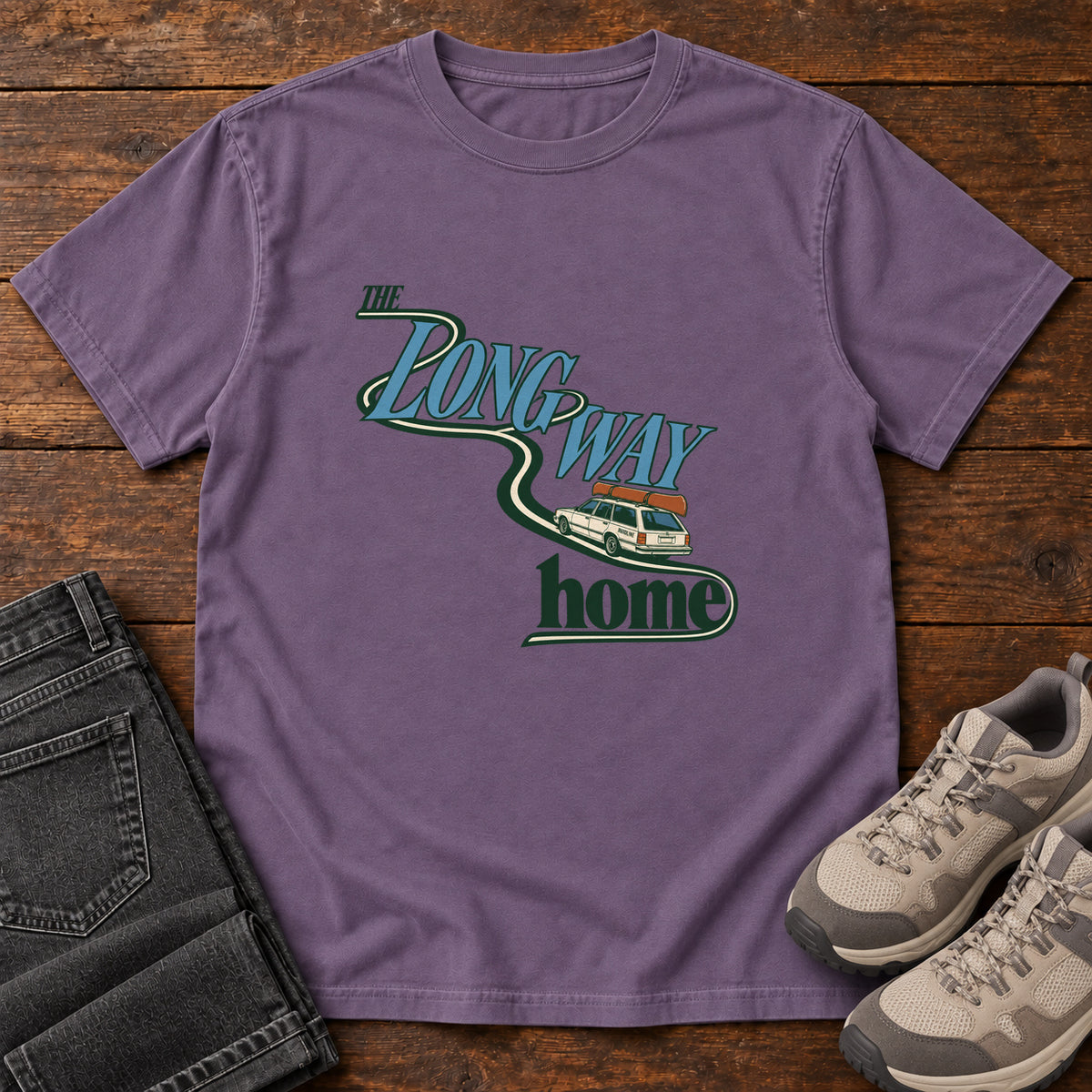 Grape The Long Way Home T-Shirt with a station wagon on a winding road woven through The Long Way Home lettering; front flat lay on wood.