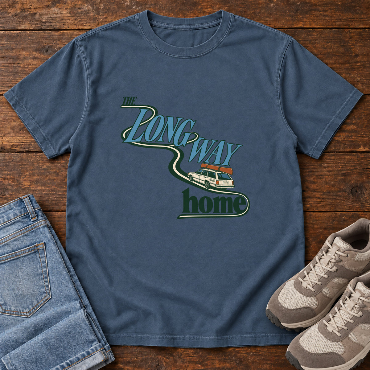 Midnight The Long Way Home T-Shirt with a station wagon on a winding road woven through The Long Way Home lettering; front flat lay on wood.