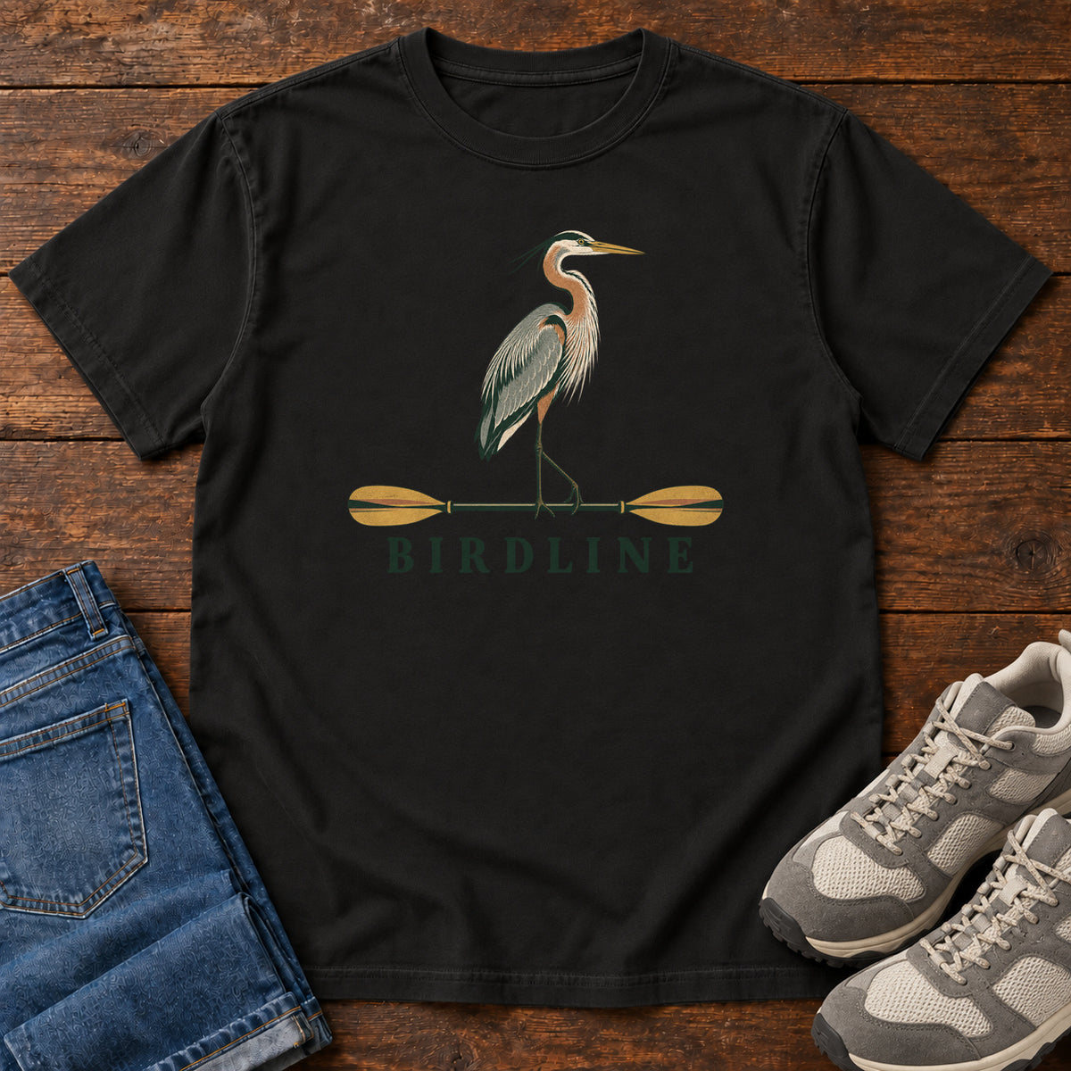 Black Great Blue Heron Paddle Birdline T-Shirt with a long-legged heron standing on a horizontal paddle above Birdline lettering; front flat lay on wood.