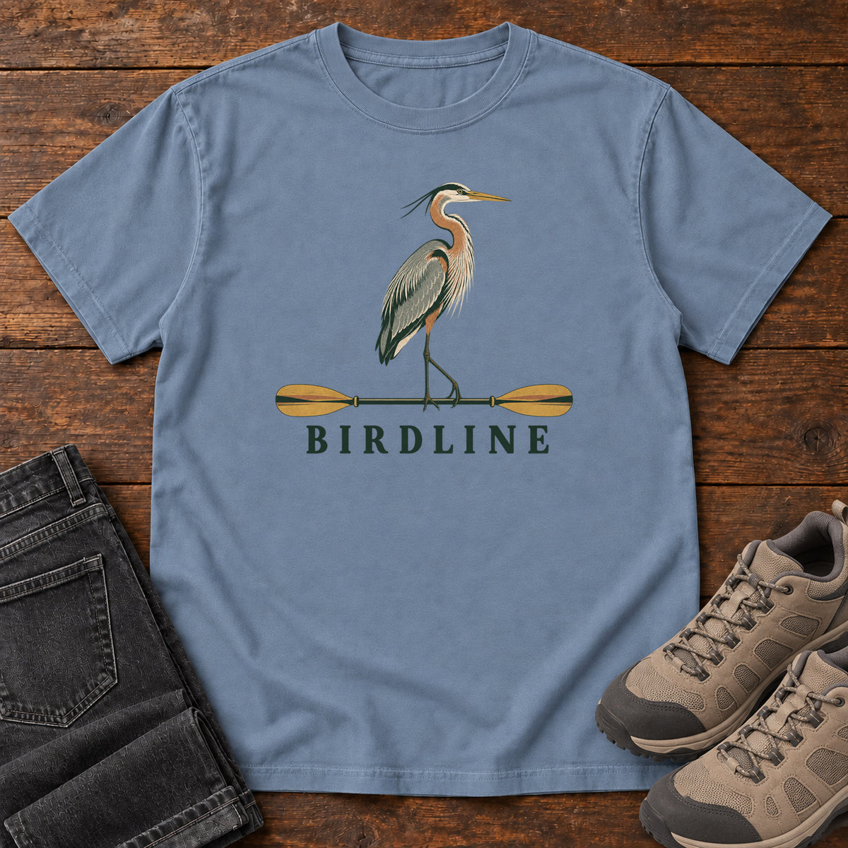 Blue Jean Great Blue Heron Paddle Birdline T-Shirt with a long-legged heron standing on a horizontal paddle above Birdline lettering; front flat lay on wood.