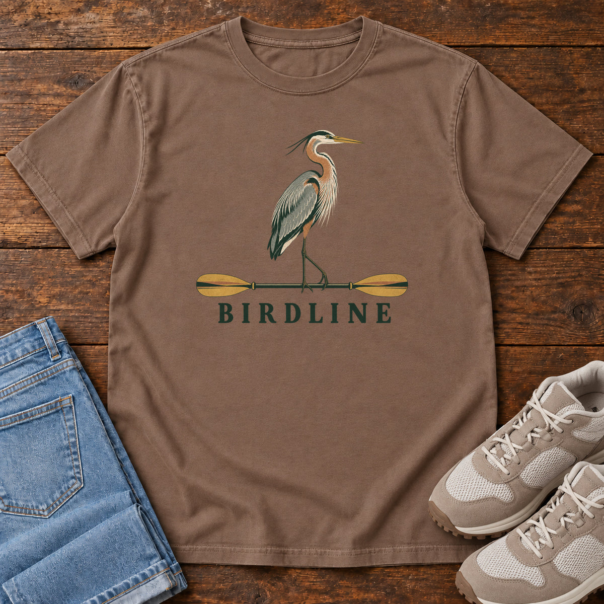 Espresso Great Blue Heron Paddle Birdline T-Shirt with a long-legged heron standing on a horizontal paddle above Birdline lettering; front flat lay on wood.