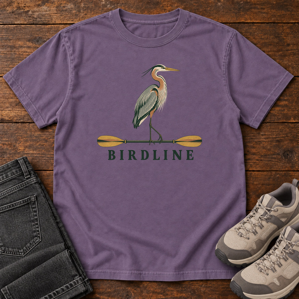 Grape Great Blue Heron Paddle Birdline T-Shirt with a long-legged heron standing on a horizontal paddle above Birdline lettering; front flat lay on wood.