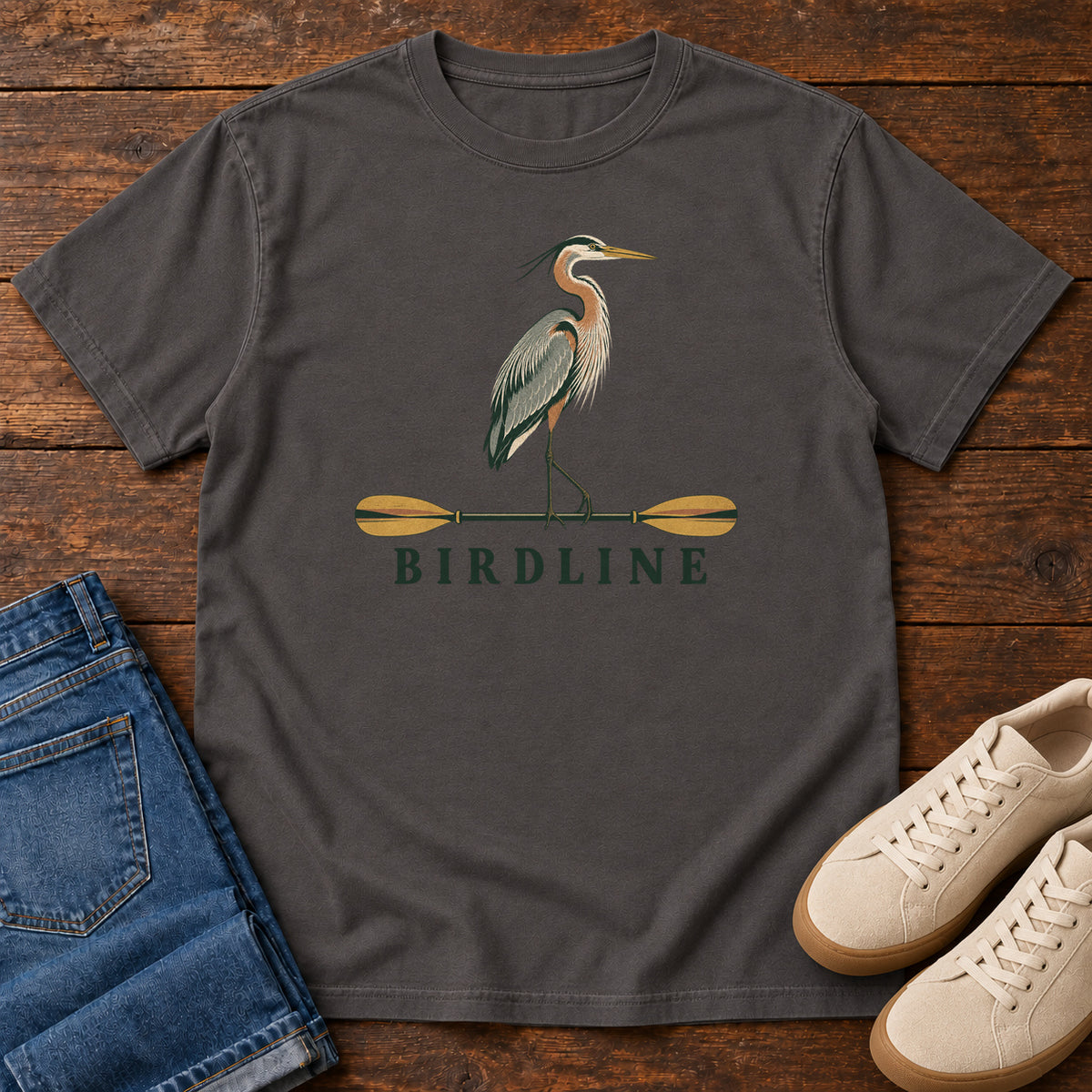 Graphite Great Blue Heron Paddle Birdline T-Shirt with a long-legged heron standing on a horizontal paddle above Birdline lettering; front flat lay on wood.