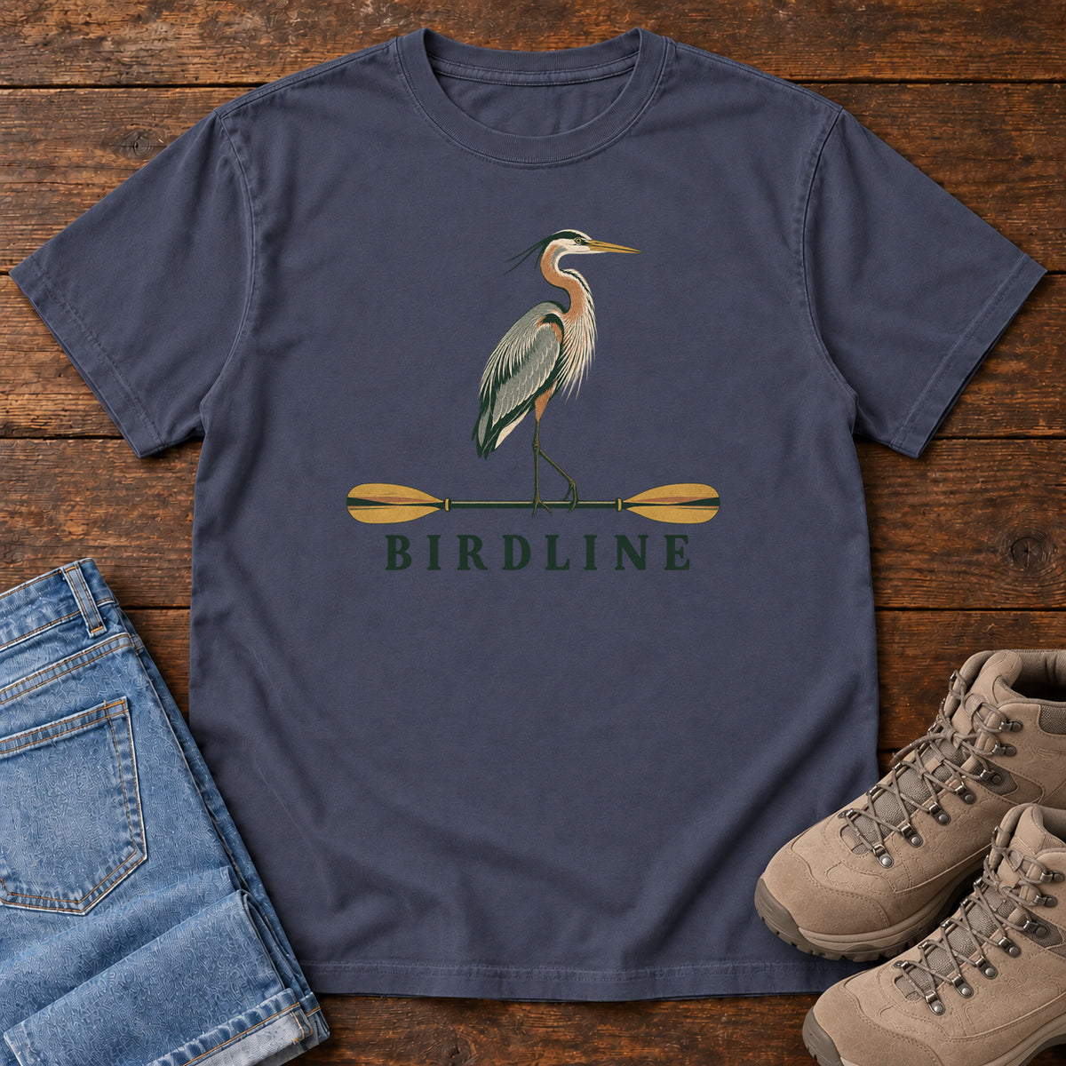 Navy Great Blue Heron Paddle Birdline T-Shirt with a long-legged heron standing on a horizontal paddle above Birdline lettering; front flat lay on wood.