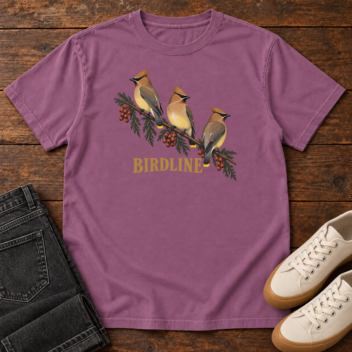 Berry Cedar Waxwings Birdline T-Shirt with three crested birds on a berry-laden evergreen branch; front flat lay on wood.