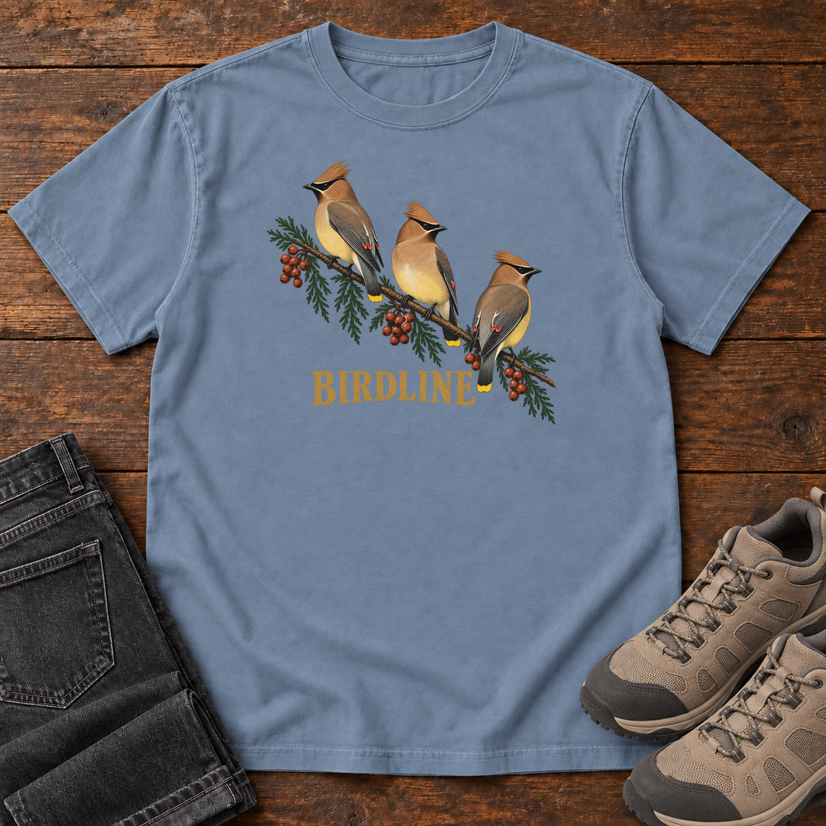 Blue Jean Cedar Waxwings Birdline T-Shirt with three crested birds on a berry-laden evergreen branch; front flat lay on wood.
