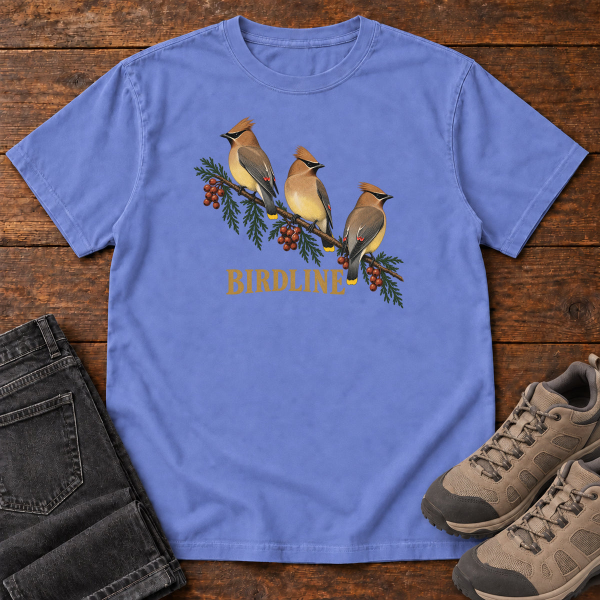 Flo Blue Cedar Waxwings Birdline T-Shirt with three crested birds on a berry-laden evergreen branch; front flat lay on wood.