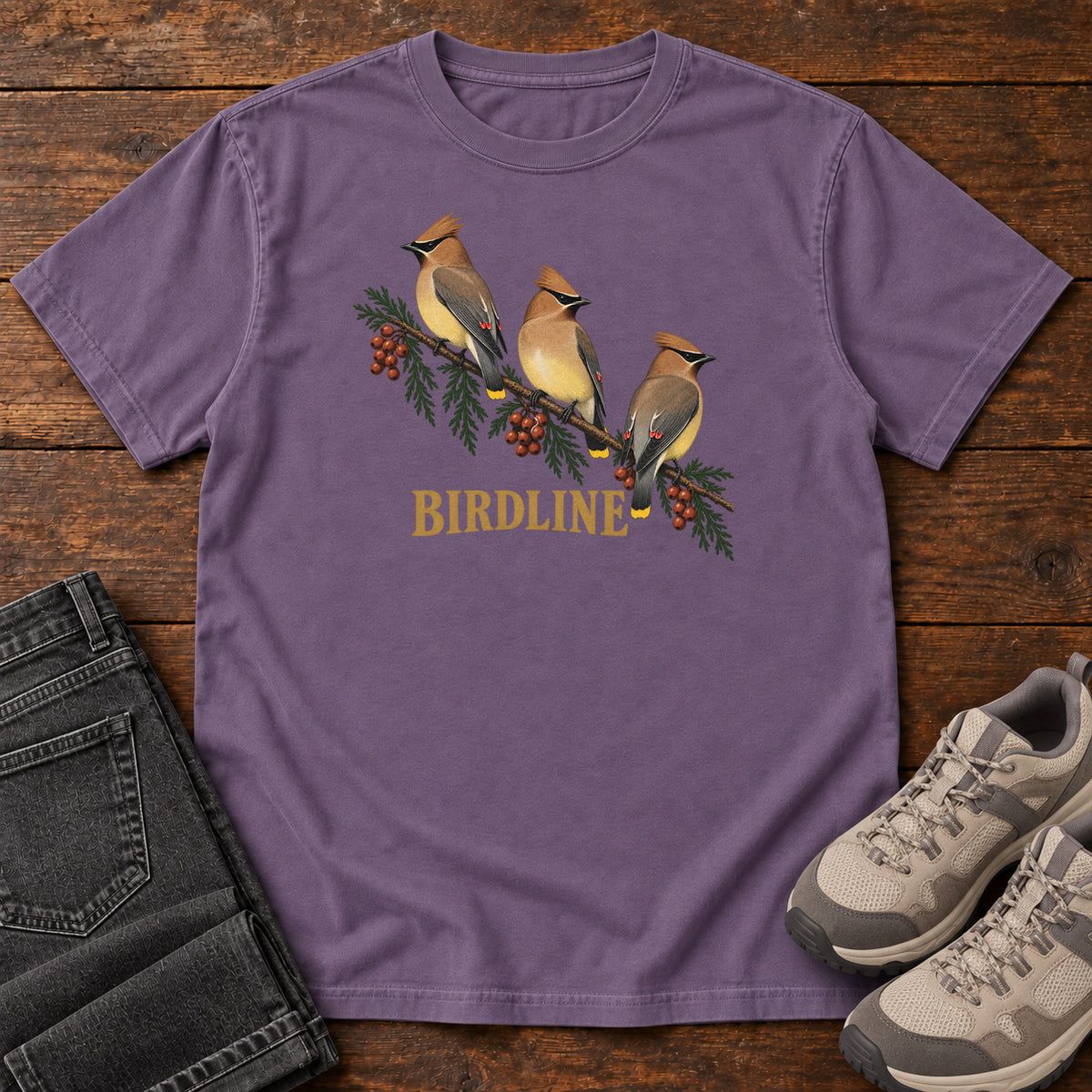 Grape Cedar Waxwings Birdline T-Shirt with three crested birds on a berry-laden evergreen branch; front flat lay on wood.