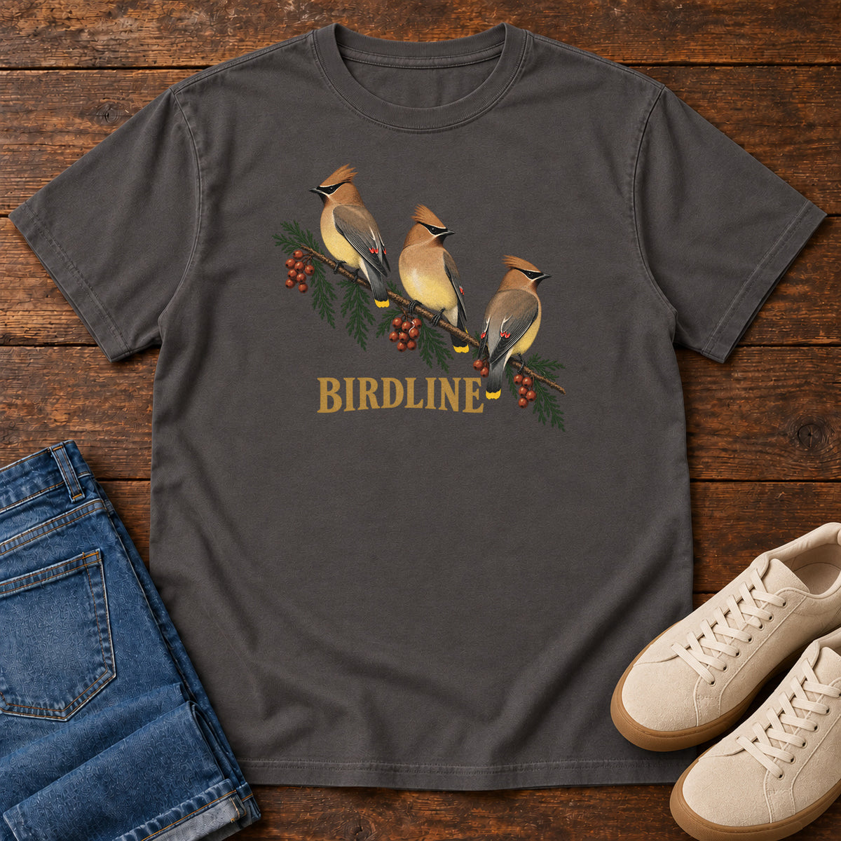 Graphite Cedar Waxwings Birdline T-Shirt with three crested birds on a berry-laden evergreen branch; front flat lay on wood.